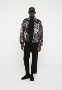 Άνδρας με μαύρο καπέλο, φλις ζακέτα με σχέδιο λύκου, μαύρο πουκάμισο, μαύρο παντελόνι και μαύρα αθλητικά παπούτσια, στέκεται μπροστά σε λευκό φόντο.