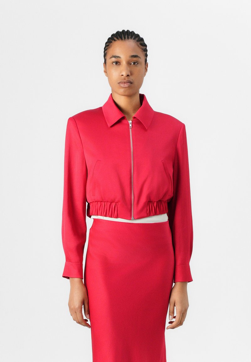 Red cropped jacket with a zip front, long sleeves, and elastic hem, paired with a matching fitted skirt. Smooth fabric, minimalist design.