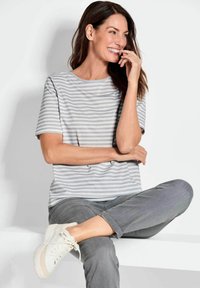 Gestreiftes grau-weißes T-Shirt mit kurzen Ärmeln und Rundhalsausschnitt, getragen mit umgeschlagenen grauen Jeans und weißen Turnschuhen, sitzt vor einem hellen Hintergrund.