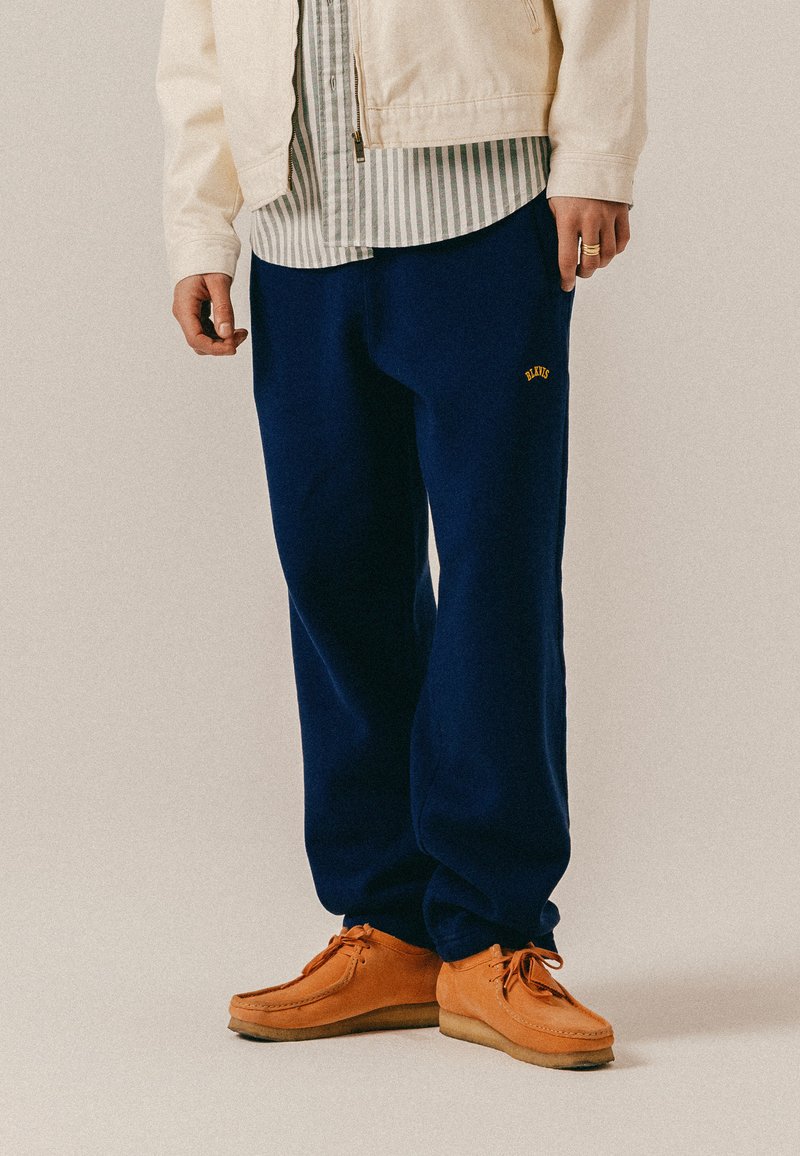 Persona che indossa pantaloni della tuta blu navy con logo giallo, maglietta a righe, giacca beige chiaro e scarpe con lacci in camoscio color cuoio.