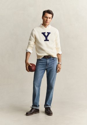 Ung mand i hvid sweater med stort Y, blå jeans og brune sko holder en fodbold mod en ensartet lys baggrund.