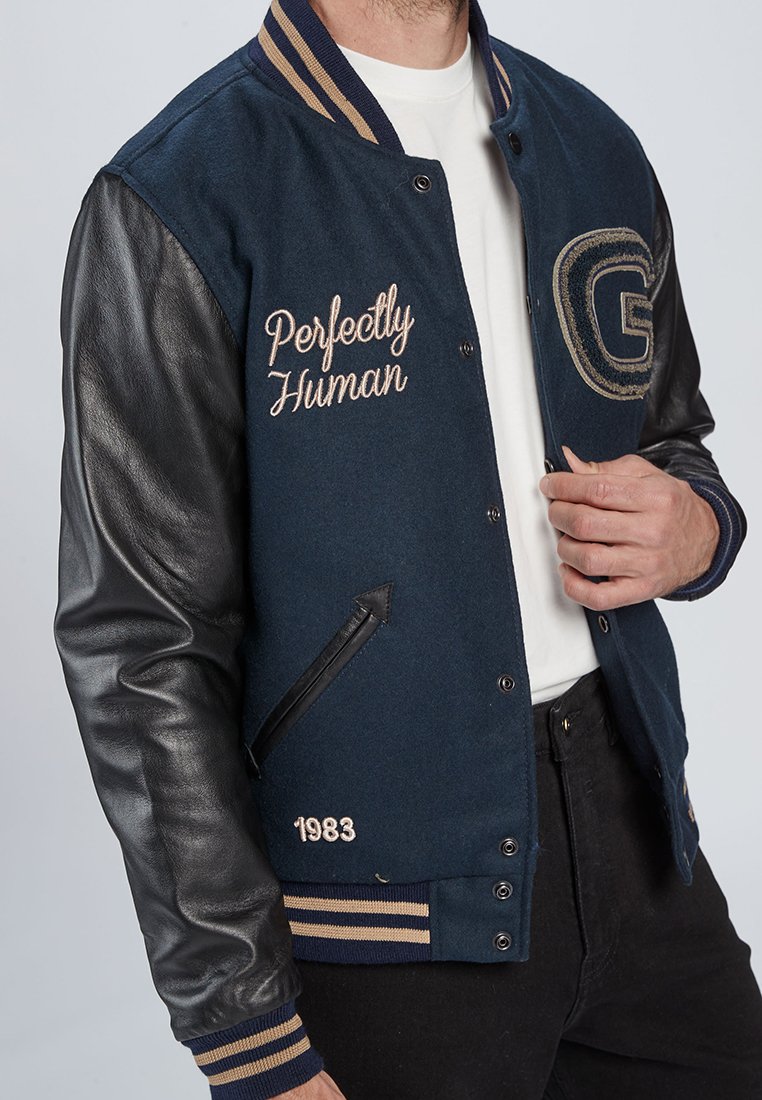 Man met een marineblauwe varsityjack met zwarte leren mouwen, een beige gestreepte kraag en manchetten, en de tekst "Perfectly Human" en "1983" geborduurd.