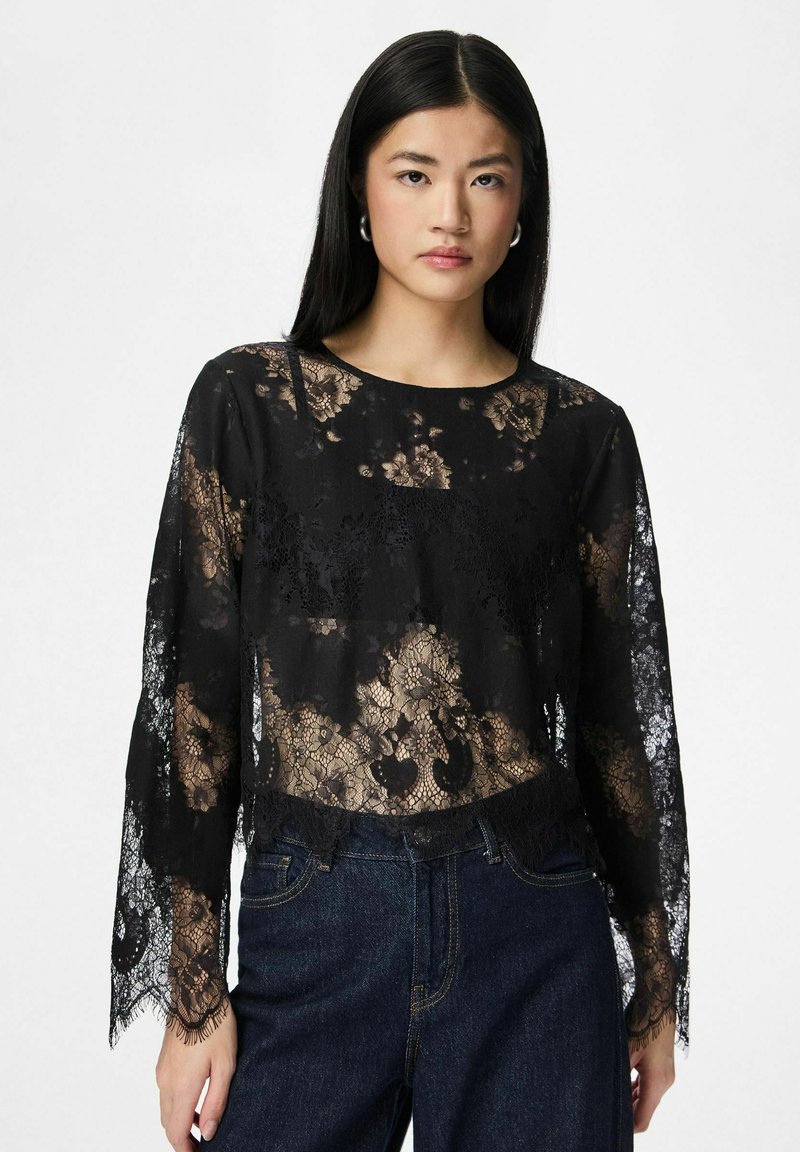 Mladá žena s dlouhými černými vlasy má na sobě černou květinovou krajkovou blůzu s dlouhým rukávem a tmavě modré džíny na jednoduchém bílém pozadí.