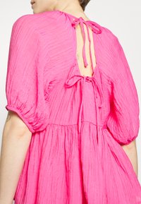Vestido rosa, texturizado, com mangas bufantes e fecho atrás com laço. Apresenta um decote profundo nas costas e um design em camadas e fluido.