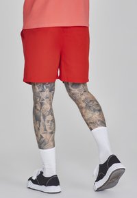 Raudoni šortai su lygiu paviršiumi, tiesia forma ir kišenėmis. Dėvimi su baltais kojiniais ir juodais sportbačiais, turinčiais detalių ant kojų tatuiruotės.