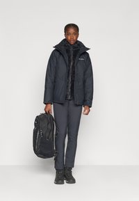 Zwarte geïsoleerde jas met capuchon, gedragen over een donkere fleece top. Grijze broek en zwarte wandelschoenen. Model houdt een zwarte rugzak vast.