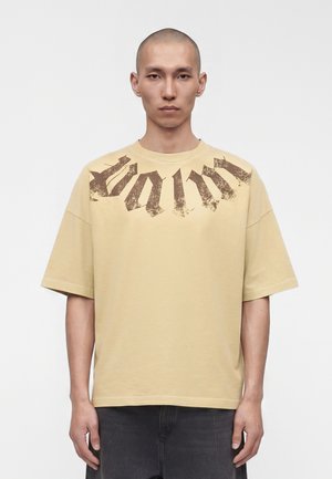 Giovane uomo con testa rasata che indossa una maglietta beige oversize con testo gotico marrone scuro sbiadito sul petto.