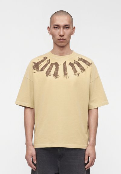 Jeune homme à la tête rasée portant un T-shirt beige oversize avec un texte gothique brun foncé délavé sur la poitrine.