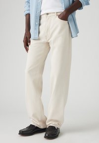 Jeans din denim bej cu picior drept, cusături proeminente, buzunare frontale și un buton la talie, asortați cu mocasini negri.