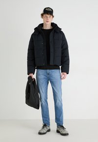 Črna puffer jakna s kapuco, ki ima pravokotne prešite sekcije. Model nosi modre kavbojke in drži črno prešito jakno. Srebrni čevlji.