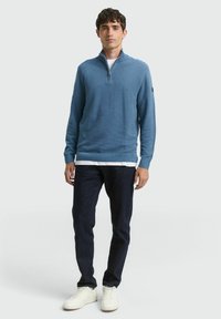 Blauer gestrickter Reißverschluss-Pullover, strukturiertes Material, lange Ärmel, gerippter Saum. Getragen über einem weißen Hemd, kombiniert mit dunklen Jeans und weißen Sneakers.