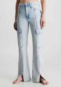 Jean de cargo de denim azul claro con cintura alta, corte acampanado, bolsillos frontales y aberturas laterales en el dobladillo. Presenta costuras visibles y botones.