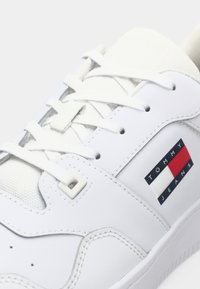 Chaussure de sport blanche avec un dessus en cuir lisse, des accents en tissu et des lacets plats. Présente un patch logo bleu, rouge et blanc portant l'inscription "TOMMY JEANS."