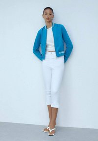 Giacca bomber blu con strisce bianche, top corto bianco, jeans bianchi a vita alta e sandali bianchi. Texture liscia e design casual.
