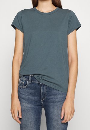 Kobieta ubrana w prosty, krótki, ciemnoturkusowy t-shirt lekko wpuszczony w niebieskie dżinsy, stojąca na białym tle.