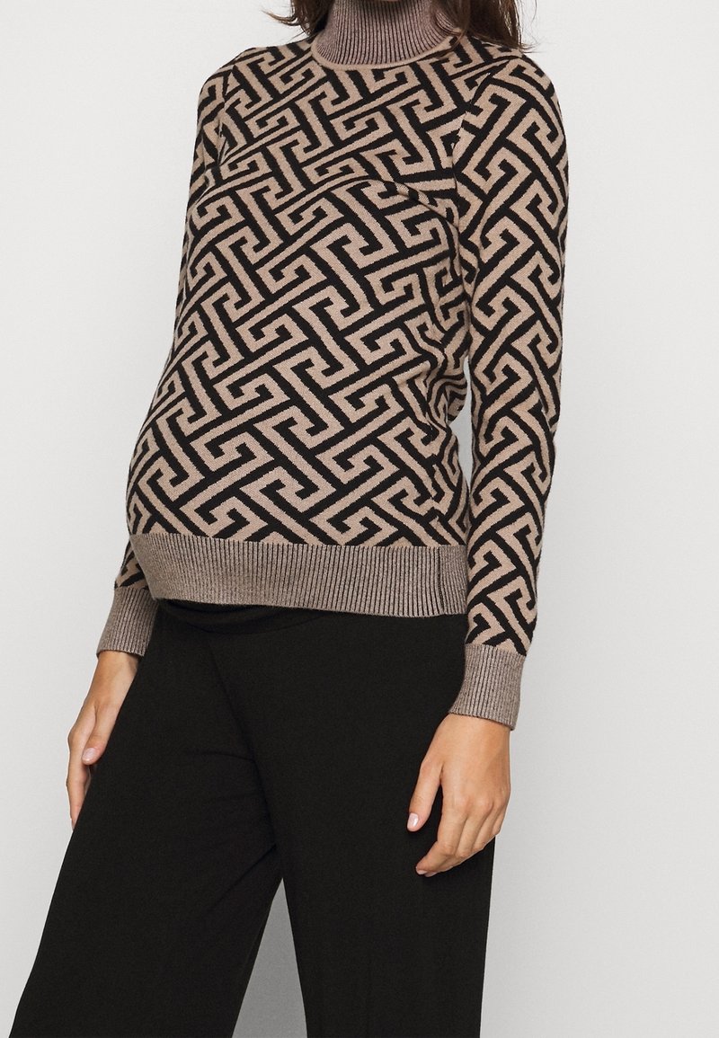 Femme portant un pull à col roulé beige et noir à motif géométrique, assorti à un pantalon noir, debout devant un fond clair uni.