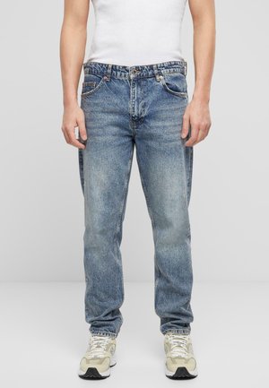 Jean en denim bleu clair avec une coupe décontractée, cinq poches et une texture délavée. Porté avec un débardeur blanc et des baskets beiges.