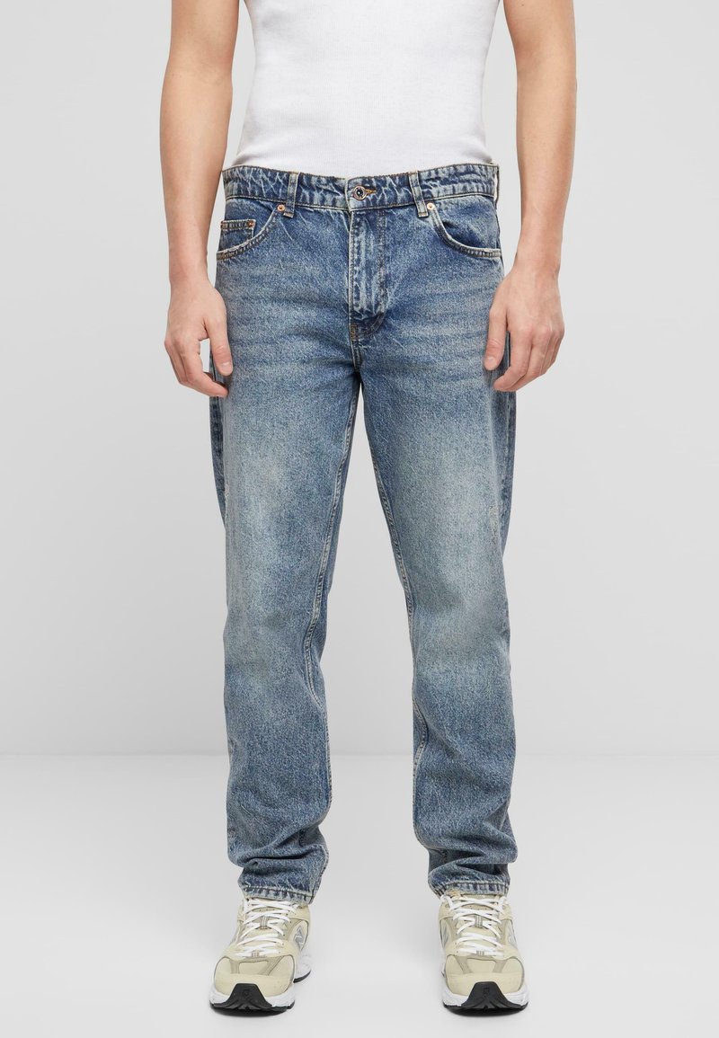Jeanși din denim de un albastru deschis, cu o croială confortabilă, cinci buzunare și o textură decolorată. Purtați cu un top alb fără mâneci și adidași bej.