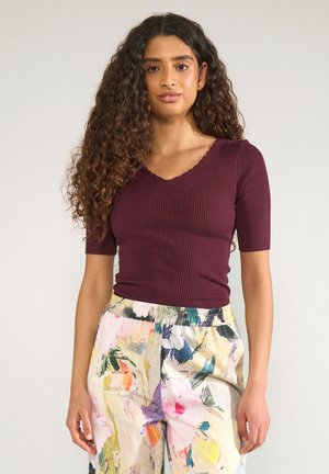 Femme aux longs cheveux bouclés portant un haut côtelé bordeaux et un pantalon coloré à motif floral, debout devant un fond uni.