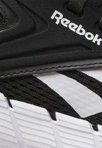 Musta urheilujalkine, jossa on teksturoitu päällinen, valkoisella Reebok-logolla ja erottuvalla musta-valkoisella raidallisella pohjasuunnittelulla.