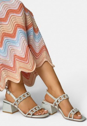 Piernas con sandalias plateadas de tacón cuadrado con tiras de cadena de pedrería, combinadas con una falda de patrón ondulado multicolor.