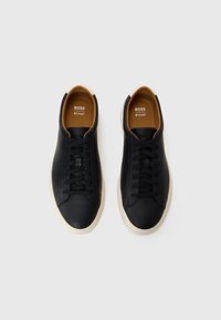 Svarte lær sneakers med en glatt tekstur, hvit gummisåle og brunt fôr. Har svarte snøringer og merkevarenavn på tungen.