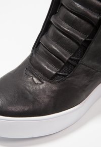 Černé kožené tenisky s vycpanou horní částí a bílou silnou gumovou podrážkou na bílém pozadí.