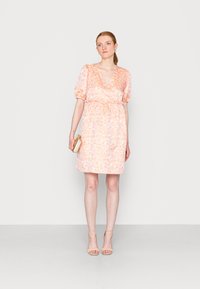 Vestido floral curto em rosa claro, com decote em V, mangas bufantes, cintura elástica e um tecido texturizado. Modelo segurando uma clutch dourada.