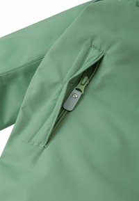Zielona kurtka z tkaniny z boczną kieszenią na zamek błyskawiczny z metalowym suwakiem i przeszyciami, prezentująca gładką, matową fakturę.