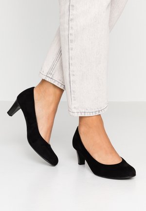 Fødder iført sorte ruskind lave hælede pumps, kombineret med lyse farvede korte jeans, stående på en glat hvid gulvflade.