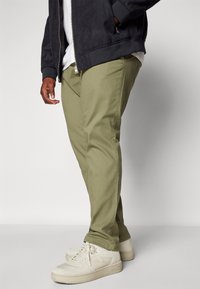 Pantalones de algodón verde oliva con un corte slim, combinados con zapatillas beige. La chaqueta es negra con un acabado texturizado y cuenta con cierre de cremallera.