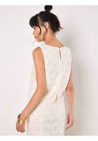 Top en dentelle avec un motif hexagonal, de couleur crème, sans manches, et une fermeture au dos en forme de clé, associé à un bracelet doré au poignet.