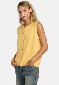 Gilet sans manches jaune avec col rond, boutons sur le devant, ourlet côtelé et texture tricotée, associé à un jean en denim taille haute.