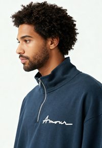 Sweat-shirt bleu marine à fermeture éclair avec un col montant, ornée du texte brodé blanc "Amour" sur la poitrine et un tissu doux et texturé.