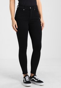 Jeans skinny de talle alto en negro, fabricados en denim, con cinco bolsillos, cremallera en la parte delantera y combinados con zapatillas negras y blancas.