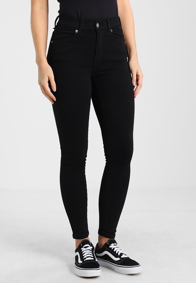 Jeans skinny de talle alto en negro, fabricados en denim, con cinco bolsillos, cremallera en la parte delantera y combinados con zapatillas negras y blancas.