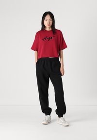 Rode cropped t-shirt met zwart geborduurd "Hugo" logo, gecombineerd met zwarte joggingbroek en lichtgrijze sneakers met witte zolen.