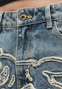 Κοντινή λήψη της μέσης μπλε τζιν με μεταλλικό κουμπί, θηλιά για ζώνη και ξεφτισμένη κεντητή λεπτομέρεια σε ξεθωριασμένο ύφασμα.