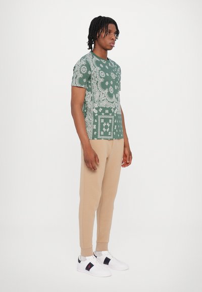 Groen T-shirt met een wit paisleypatroon, gecombineerd met beige sweatpants en witte sneakers met marineblauwe accenten.