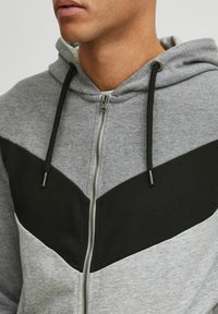 Gray zip-up hoodie with black chevron design, adjustable black drawstrings, and textured fabric. Features a metal zipper and casual fit.