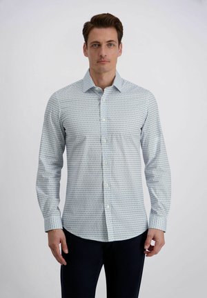 Uomo con camicia a maniche lunghe azzurra con motivi e pantaloni scuri, in piedi davanti a uno sfondo grigio chiaro uniforme.