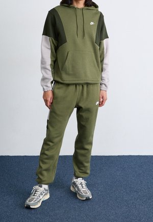 Person trägt olivgrünen und grauen Nike-Hoodie sowie passende Jogginghose mit grauen und weißen Sneakers, steht auf blauem Teppich.