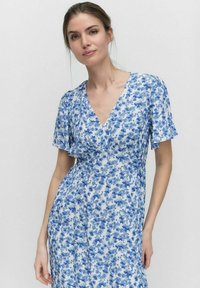 Kukallinen mekko, jossa on valkoinen tausta sinisillä kukkakuviolla, lyhyet lepattelevat hihat ja V-pääntie, valmistettu kevyestä kankaasta.