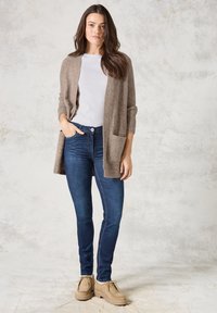 Cardigan en maille brun clair avec une ouverture sur le devant et deux poches, associé à un jean skinny bleu foncé et des chaussures à lacets beige, sur fond neutre.