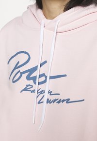 Ljusrosa hoodie med dragsko, med en framträdande logo i marinblått tryck. Mjuk tygtextur, avslappnad passform och ribbade ärmslut.