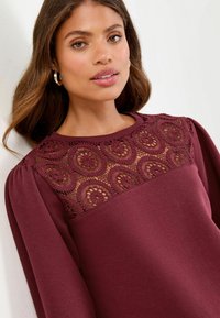 Bordo majica s dugim rukavima s čipkastim detaljem na prsima s cvjetnim uzorcima i teksturiranim detaljima. Mekana tkanina s napuhanim rukavima.