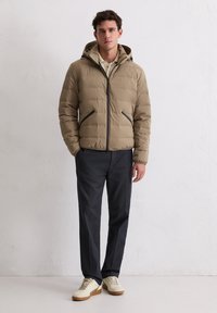 Beige gesteppte Jacke mit Reißverschluss und Kapuze, mit schwarzen Akzenten. Getragen über einem hellen Hemd mit dunkler, strukturierter Hose und cremefarbenen Turnschuhen.
