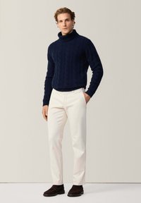 Marine kabelgebreide coltrui, witte slim-fit broek en donkerbruin suède schoenen. Eenvoudig ontwerp, gestructureerde stof en strakke lijnen.