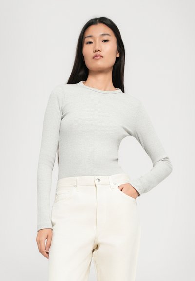Mlada ženska s dolgimi temnimi lasmi nosi svetlo sivo rebrasto top z dolgimi rokavi in hlače v svetli off-white barvi z visokim pasom, pozira z eno roko v žepu.