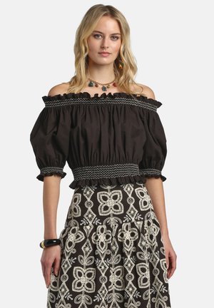 Blonde vrouw gekleed in een zwart off-shoulder blouse met opgeblazen mouwen en een zwarte rok met grote witte bloemrijke borduursels.